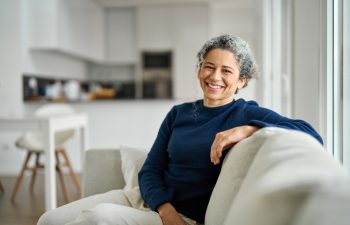 Happy middle aged woman sitting on comfy sofa in living room at home laughing. Smiling mature older lady relaxing on couch looking at camera on couch in modern cozy house. Portrait.
