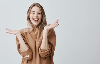 Pretty beautiful woman with blonde long hair looking at camera having excited and happy facial expression, clapping with her hands against blank studio wall, expressing her excitement with present